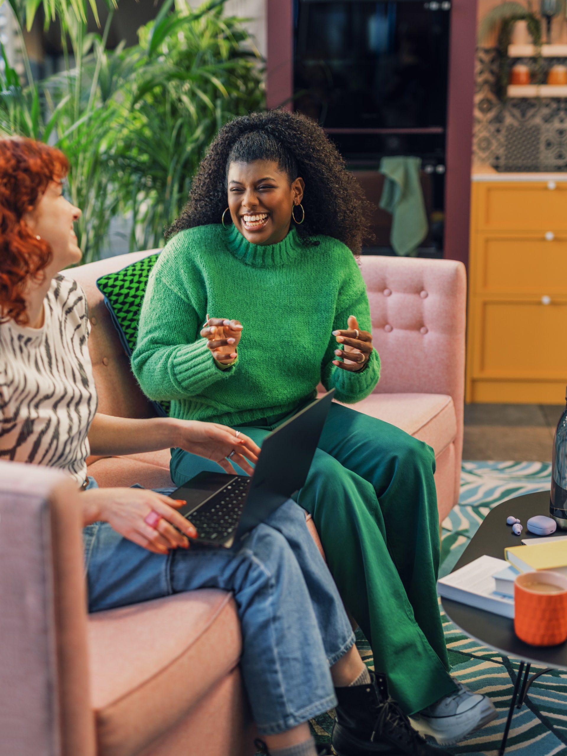 A Black woman wearing a green sweater and green pants smiling and talking to a woman with red hair holding a laptop