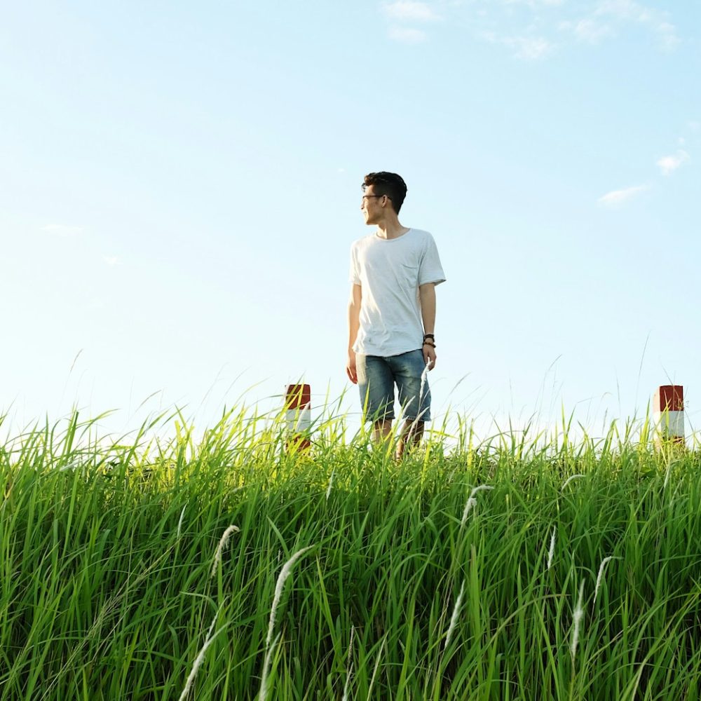 Man wearing white t-shirt and blue jean shorts look to his side over a field of grass on a bright sunny day
