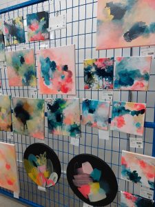An array of abstract art featuring pink, blue, and yellow hues hung on a blue metal backing at Miranda's art show.