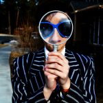 Man in a striped suit wearing sunglasses holds a magnifying glass in front of his face.